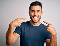 Man in blue t-shirt with dark hair pointing to his smile with both pointer fingers 