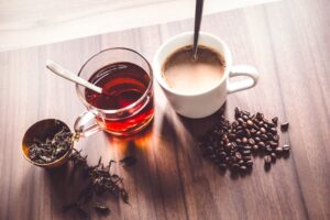 Coffee beans and tea leaves next to cups of tea and coffee on a wooden surface